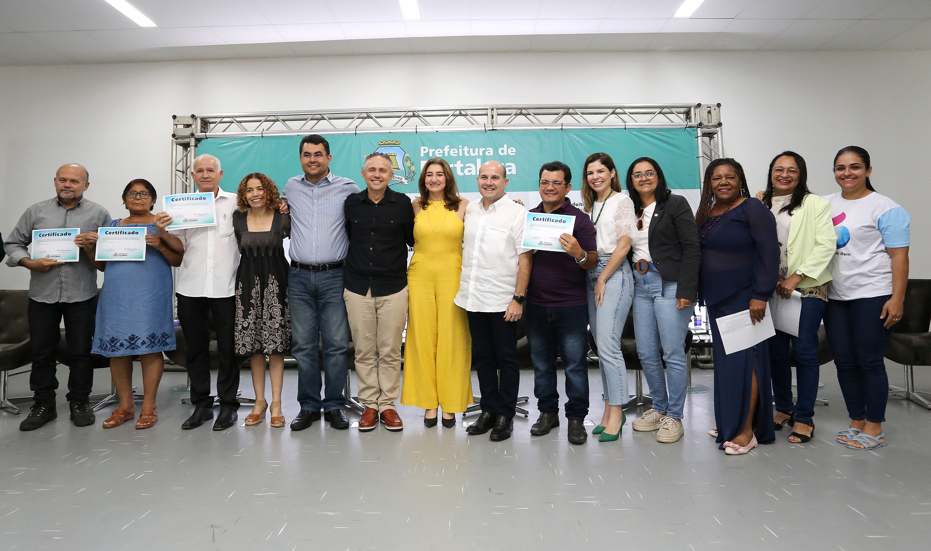 prefeito e pessoas em pé em cima de palco segurando documento e sorrindo para a foto